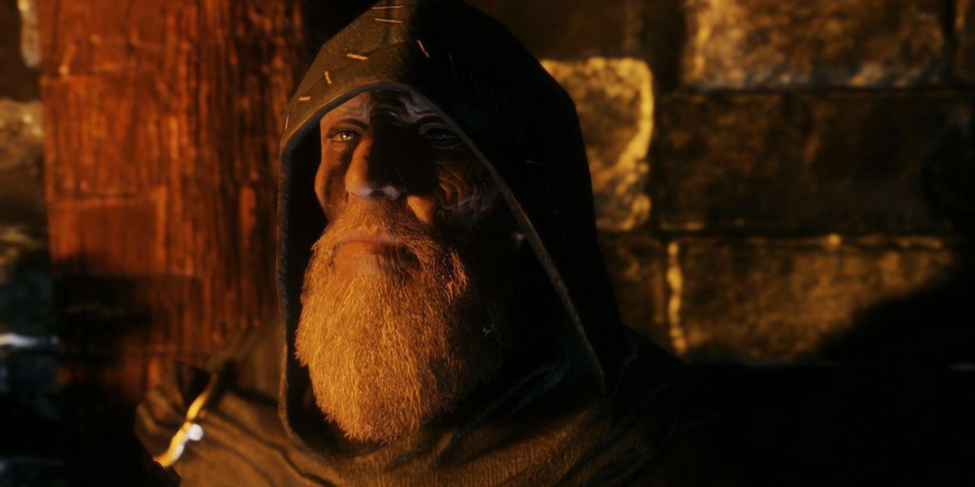 An old wizard looks at the camera in a room lit by a nearby fire.