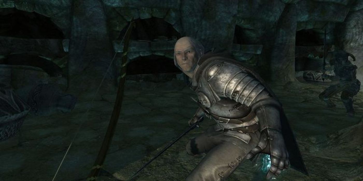 A balding man dressed in metal armor crouches inside a dark cave with blue magic pulsing in his palm.