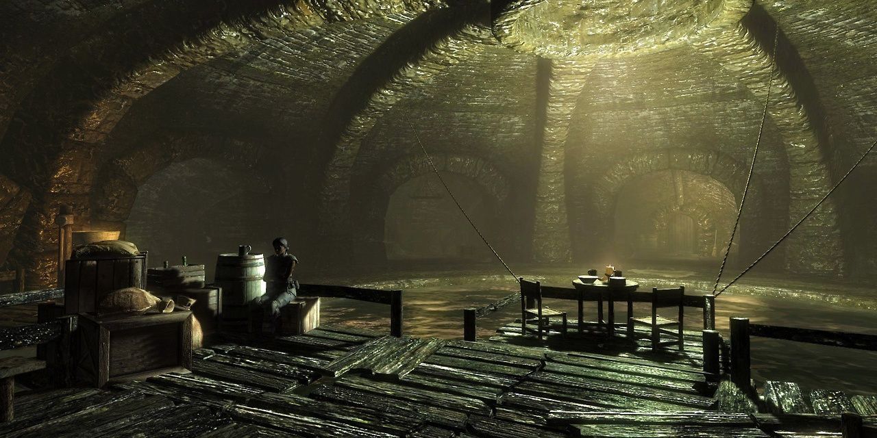 A screenshot of the entrance of the Thieves' Guild in Skyrim. Tonilia sits on a bench near barrels and crates, while light streams through a hole in the wall, reflecting off the dungeon's water.