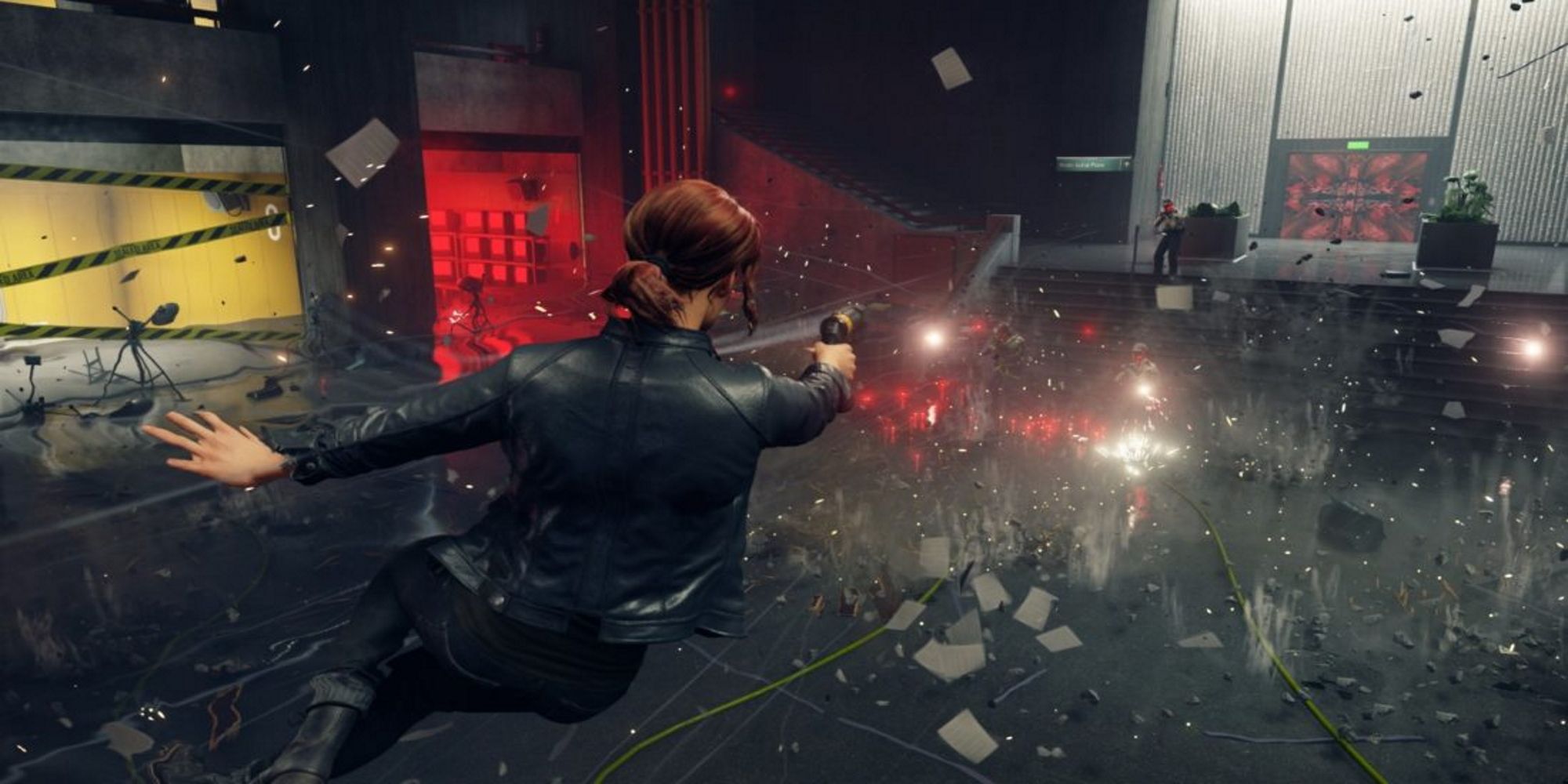 Jesse Faden from Control suspended in the air as she fires her service weapon at enemies, an explosion in the area.