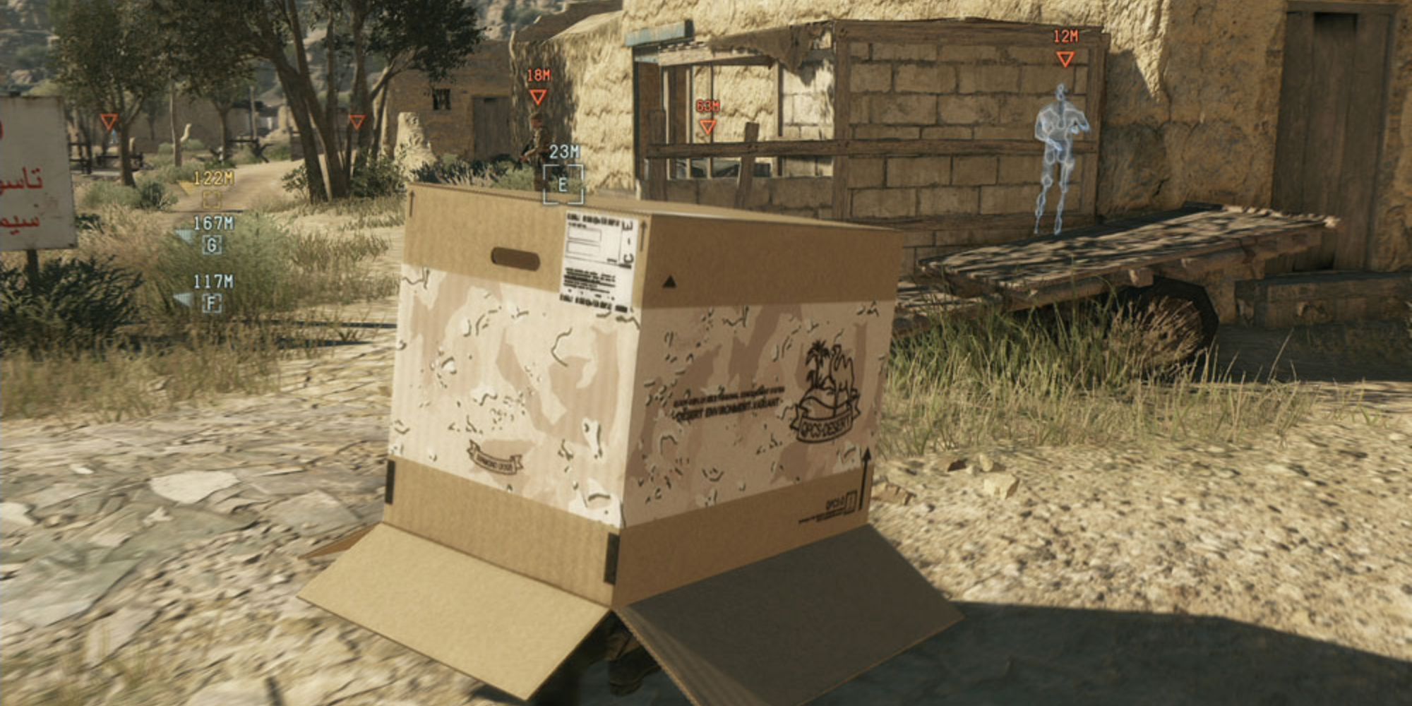 Fast Travel In Video Games a mid shot of Snake hiding beneath a cardboard box in Metal Gear Solid 5 The Phantom Pain on a sand-covered road with tufts of grass near him and buildings, trees and enemies off in the distance