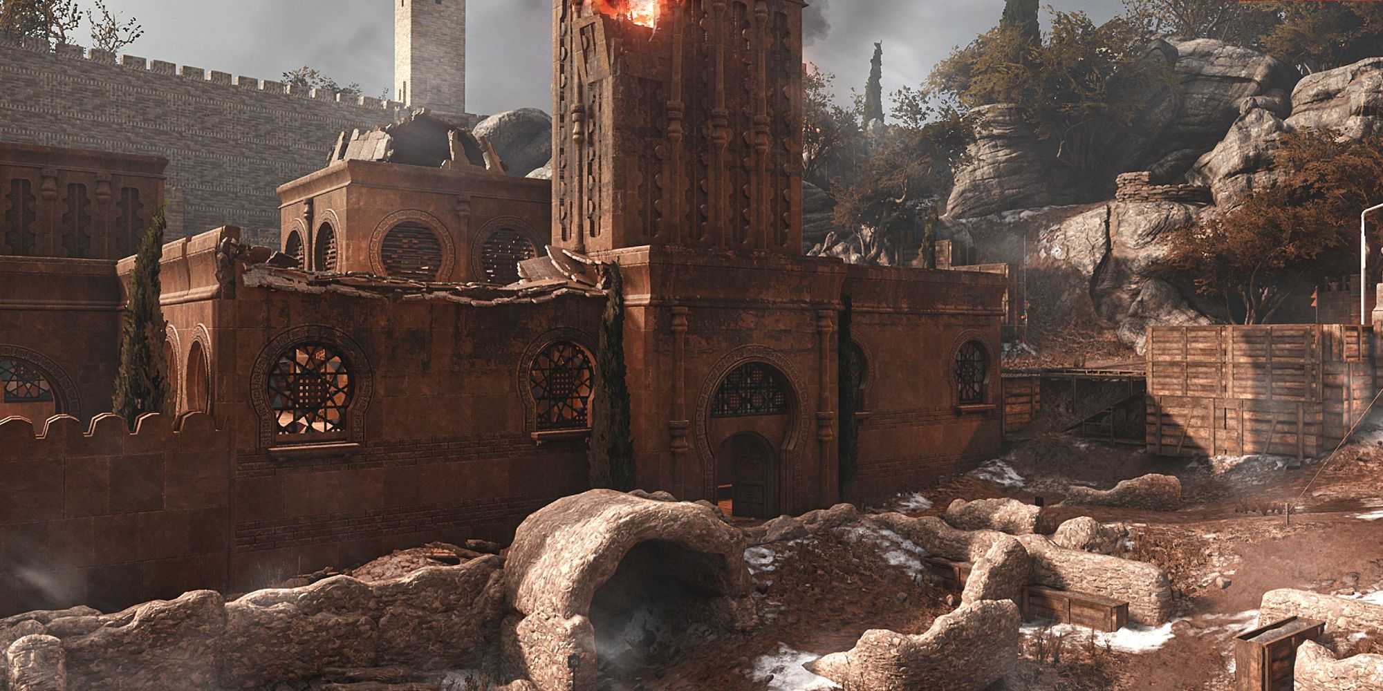 Insurgency Sandstorm Maps a mid shot of the map Citadel with a red-brick ornate building on fire in the centre against a towering stone wall in the background 