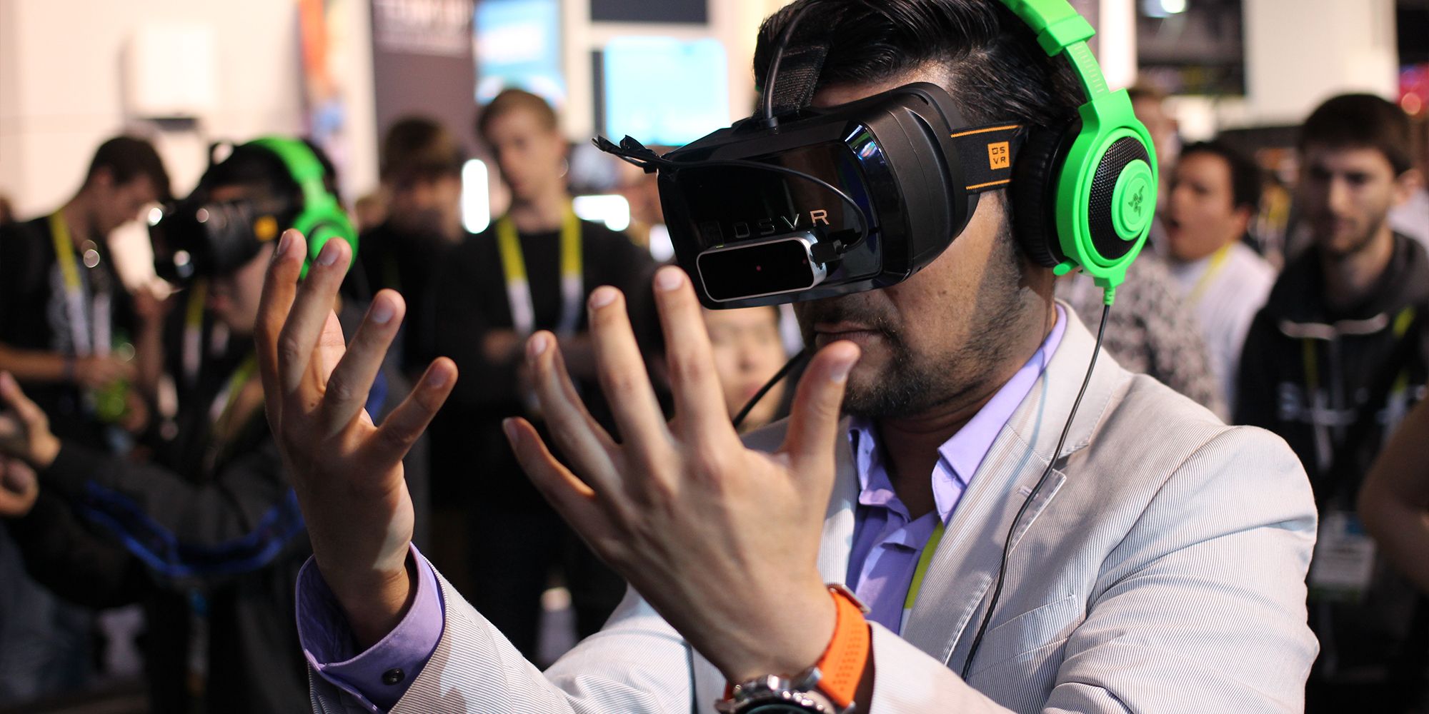 Man holds his hands out in front of his face while wearing VR headset among crowd of onlookers, beside another man in a VR headset