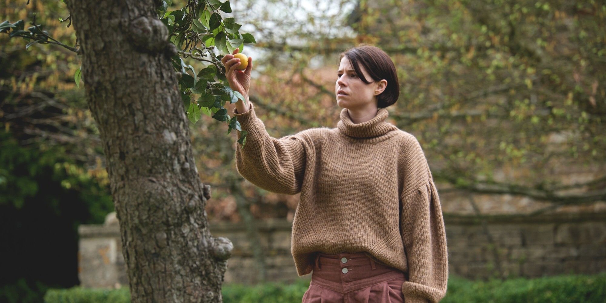 Jessie Buckley as Harper in Men plucks an apple from a tree.