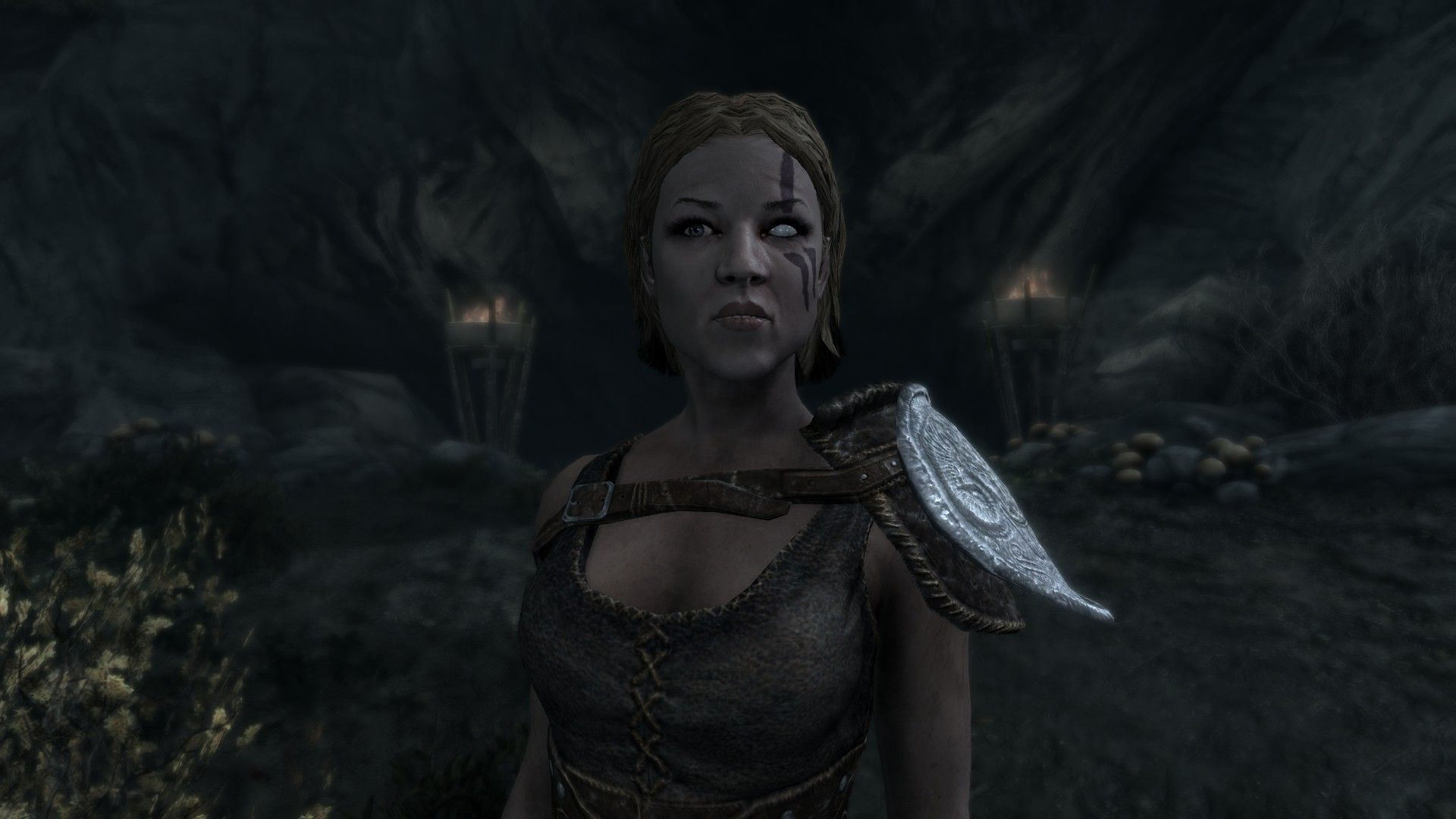 A woman dressed in light armor and warpaint stands outside a cave lit by two braziers.