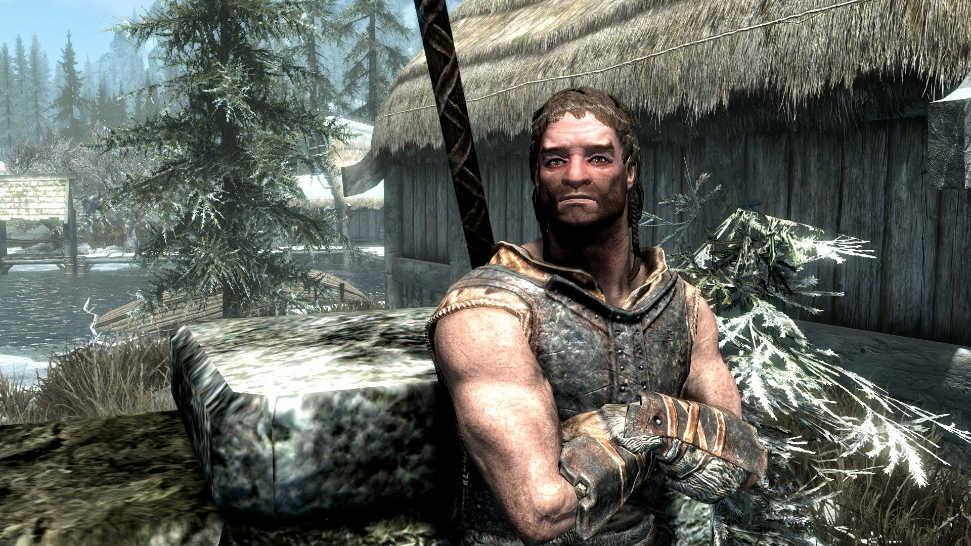A muscular man with close-cropped hair leans against a stone fence with his arms crossed in an old town.