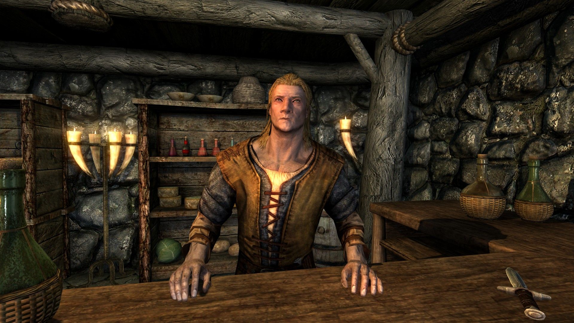 A shopkeeper leans on his counter with a variety of food, potions and weapons laying about the store.