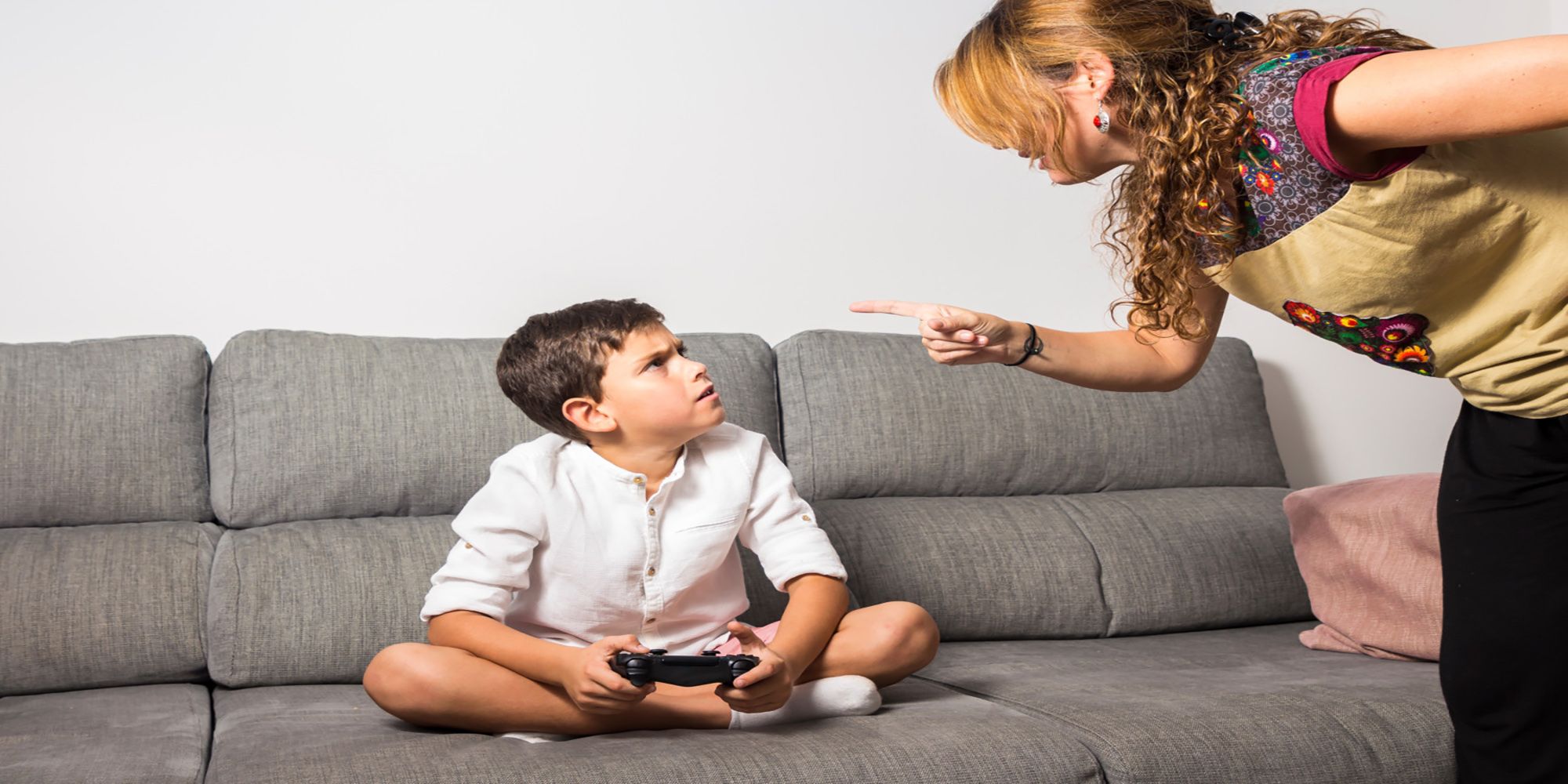 A mother yells at her son, who is sitting on a couch trying to play a video game.