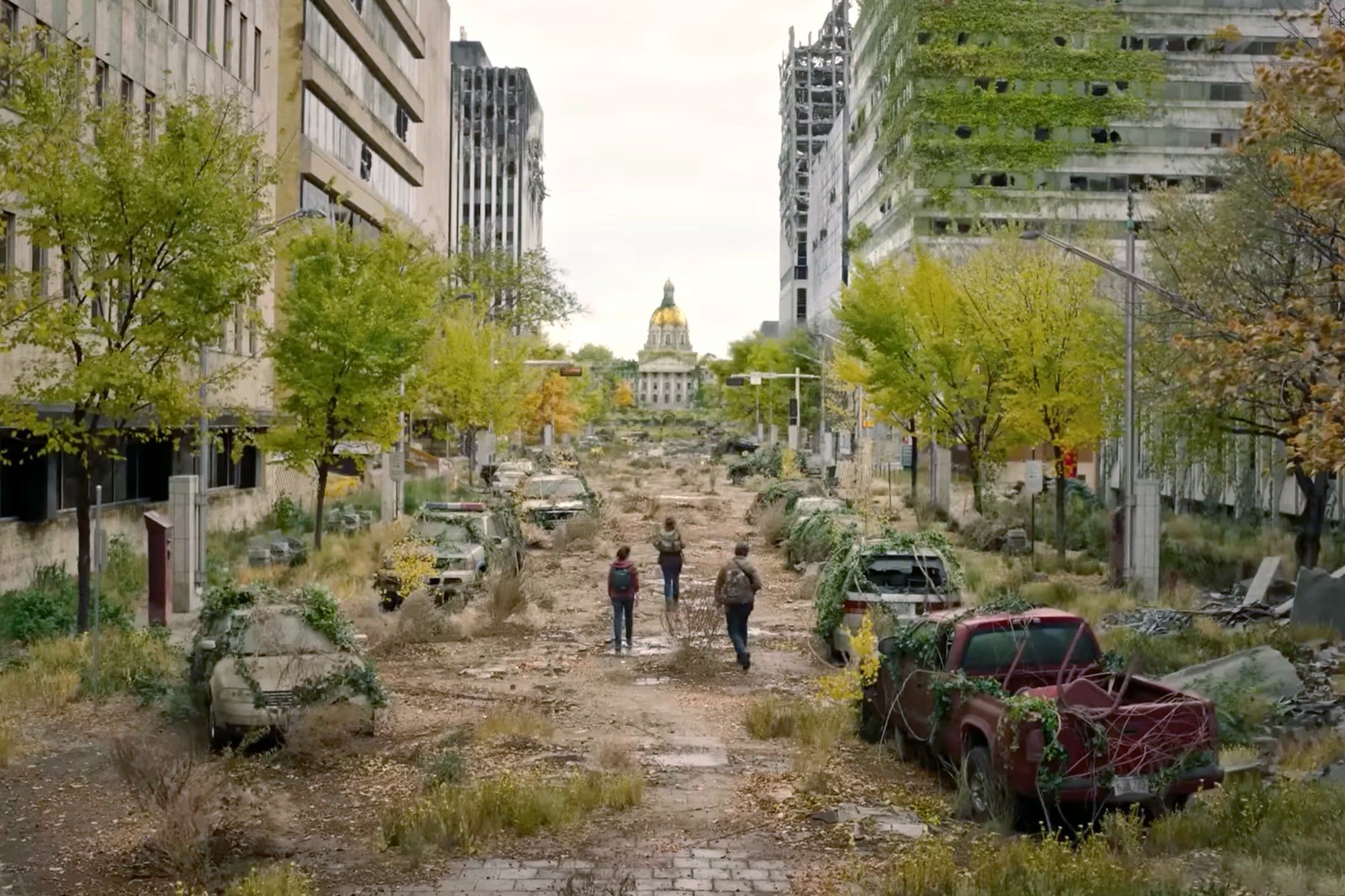 An image from a set from The Last of Us showing a rundown street with trees and grass growing in the road and abandoned cars and the three main characters in the show in the middle of the road
