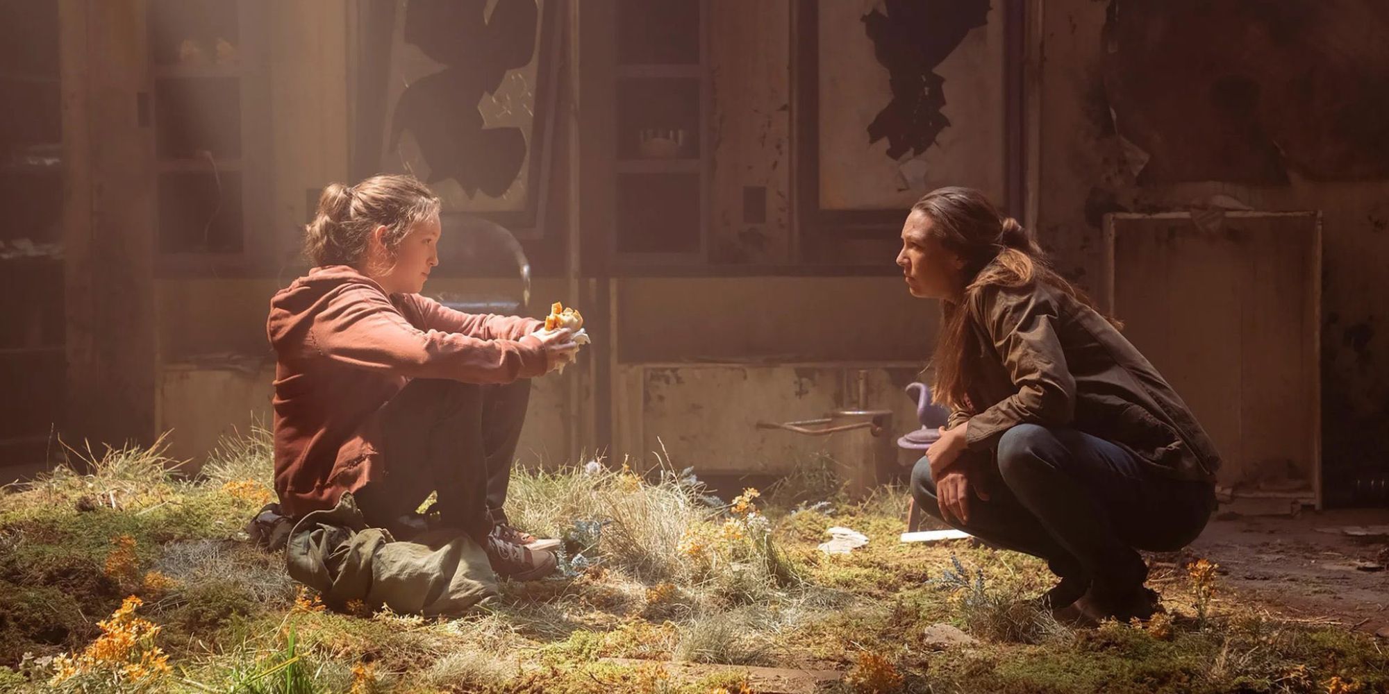 Image showing Ellie and Tess in The Last Of Us TV show with Ellie sitting down on grass lit by overhead sun inside a building while Tess squats to talk to her