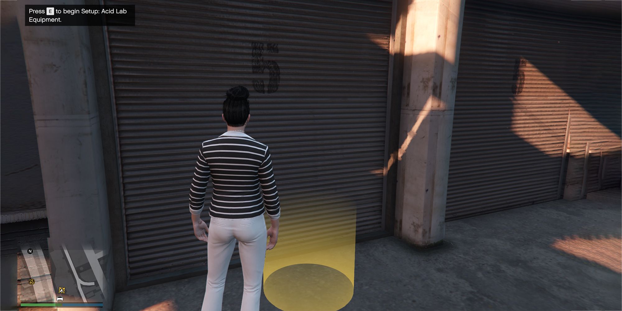 The image shows a Grand Theft Auto Online player standing outside a warehouse next to a circular yellow marker on the ground.  The text in the upper left reads 'Press E to begin setting up the acid lab equipment'.