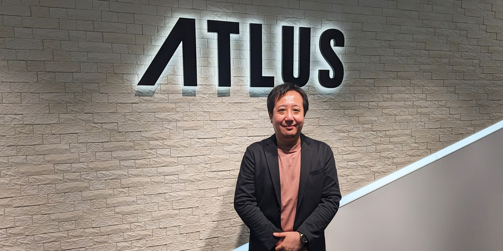 Persona 5 Tactica producer Atsushi Nomura in front of an Atlus sign.