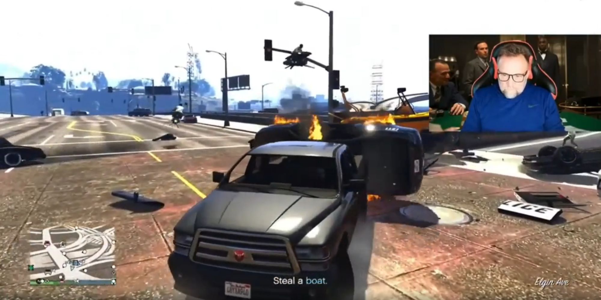 A truck surrounded by burning vehicles with GTA 5 actor Ned Luke on a screen in the top right corner