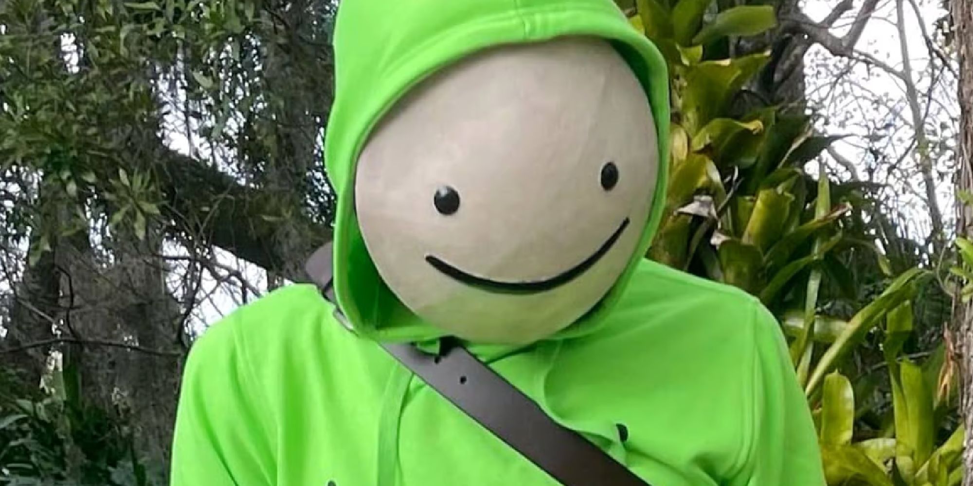Dream YouTuber standing in front of trees in a lime green hoodie with his iconic white circular mask with a smiley face painted on top