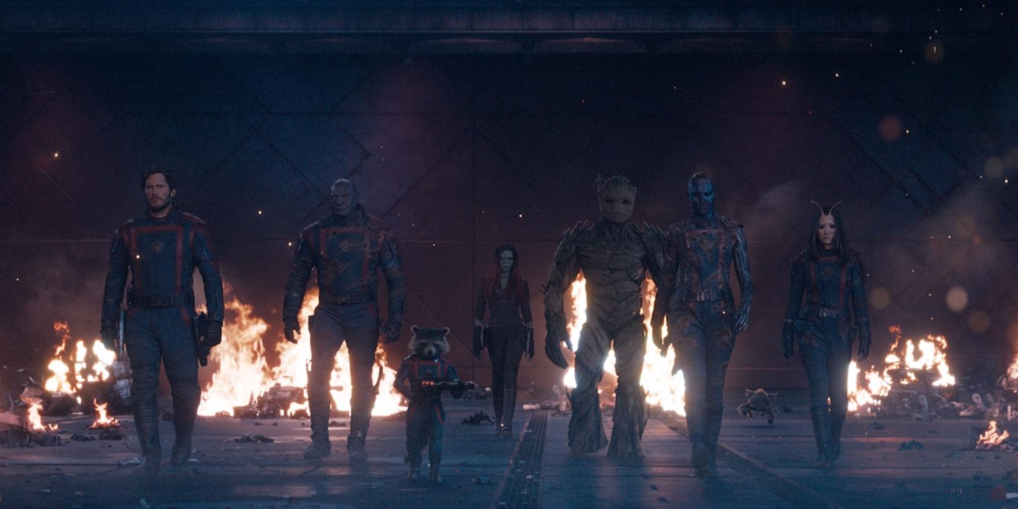 Star-Lord, Drax, Rocket, Gamora, Groot, Nebula, and Mantis walking toward the camera as creatures flee behind them and a fire burns.
