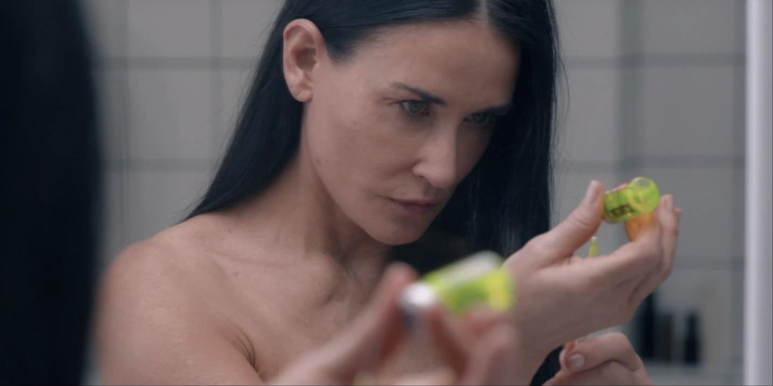 Demi Moore's character through a bathroom mirror about to inject herself with a green serum in The Substance.