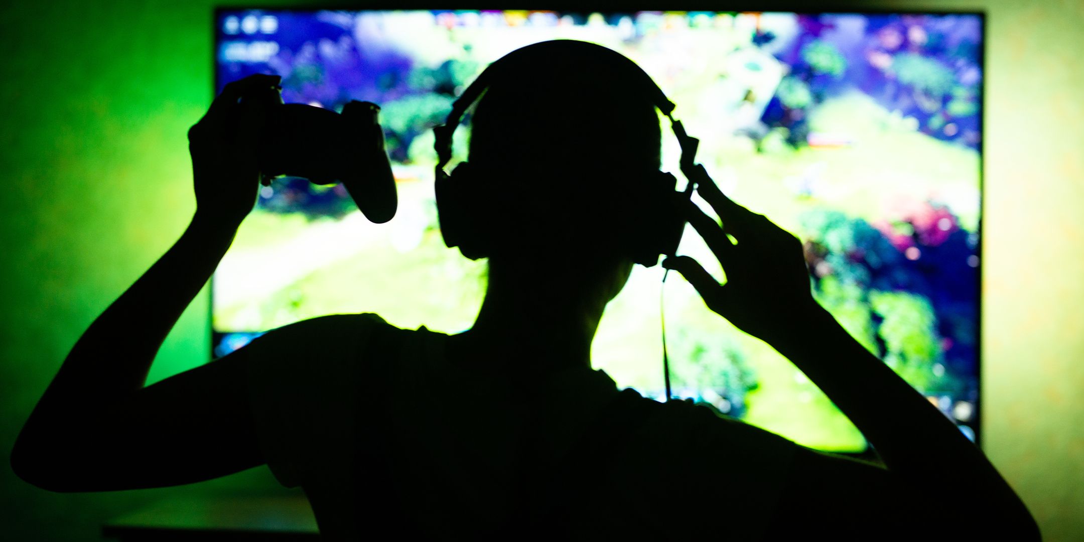 Silhouette of a gamer in a green lit room holding up a controller in front of a blurred TV screen.