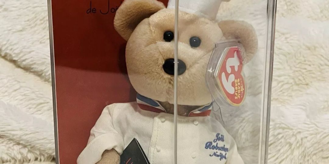 A tan teddybear in a chef's costume sits in a glass case
