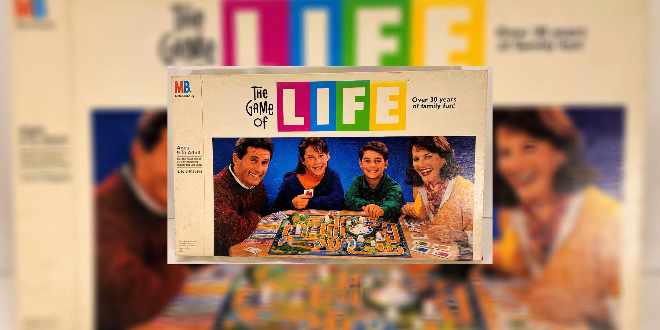 The game cover for The Game of Life depicts a family of four smiling at the camera while in front of the game board and all its pieces.