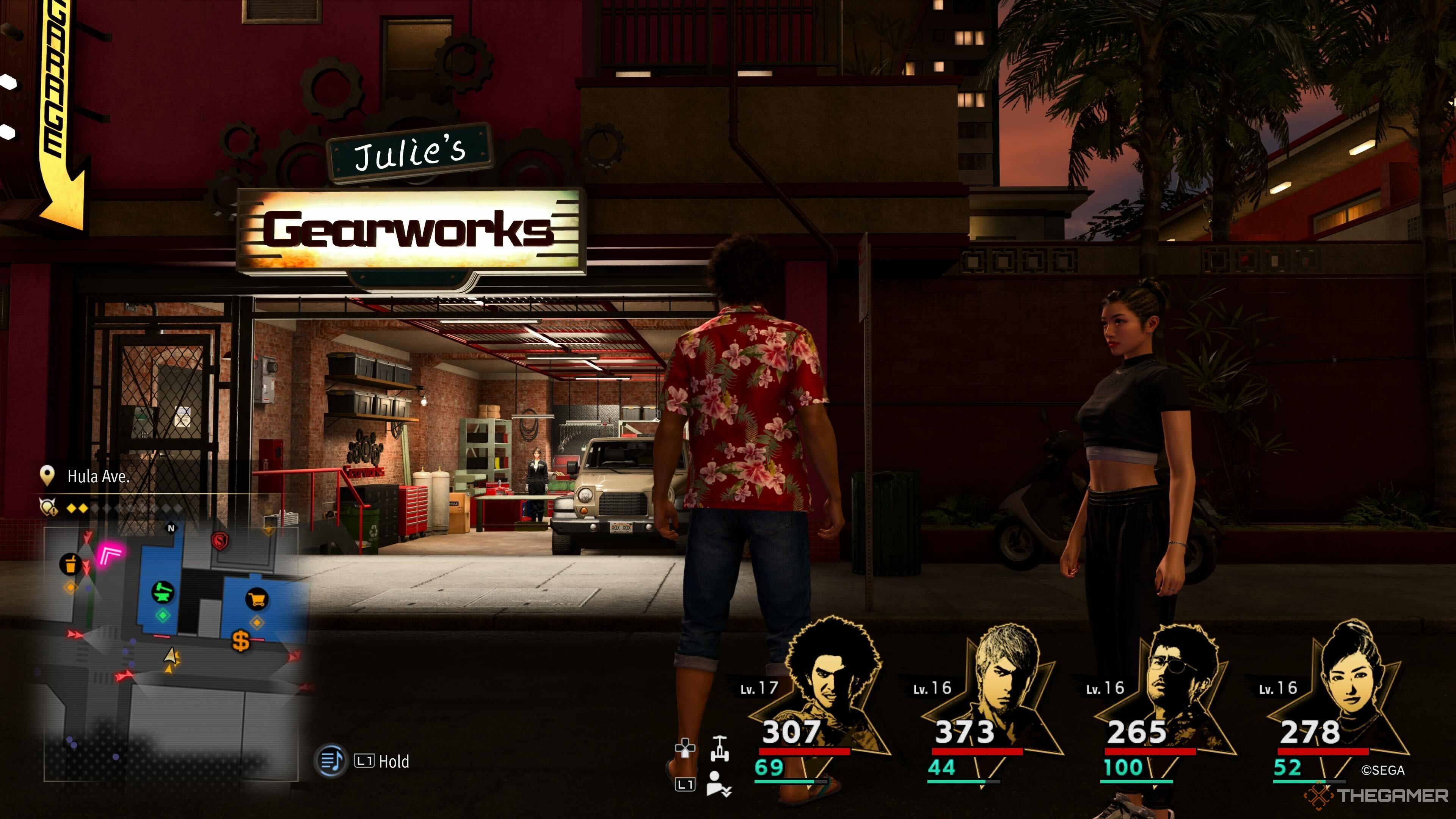 Ein Screenshot aus Like a Dragon: Infinite Wealth, der Ichiban und Chitose vor Julies Gearworks zeigt, mit einem Lastwagen in der Garage und Julie, die hinter einem klappbaren Tisch steht. 