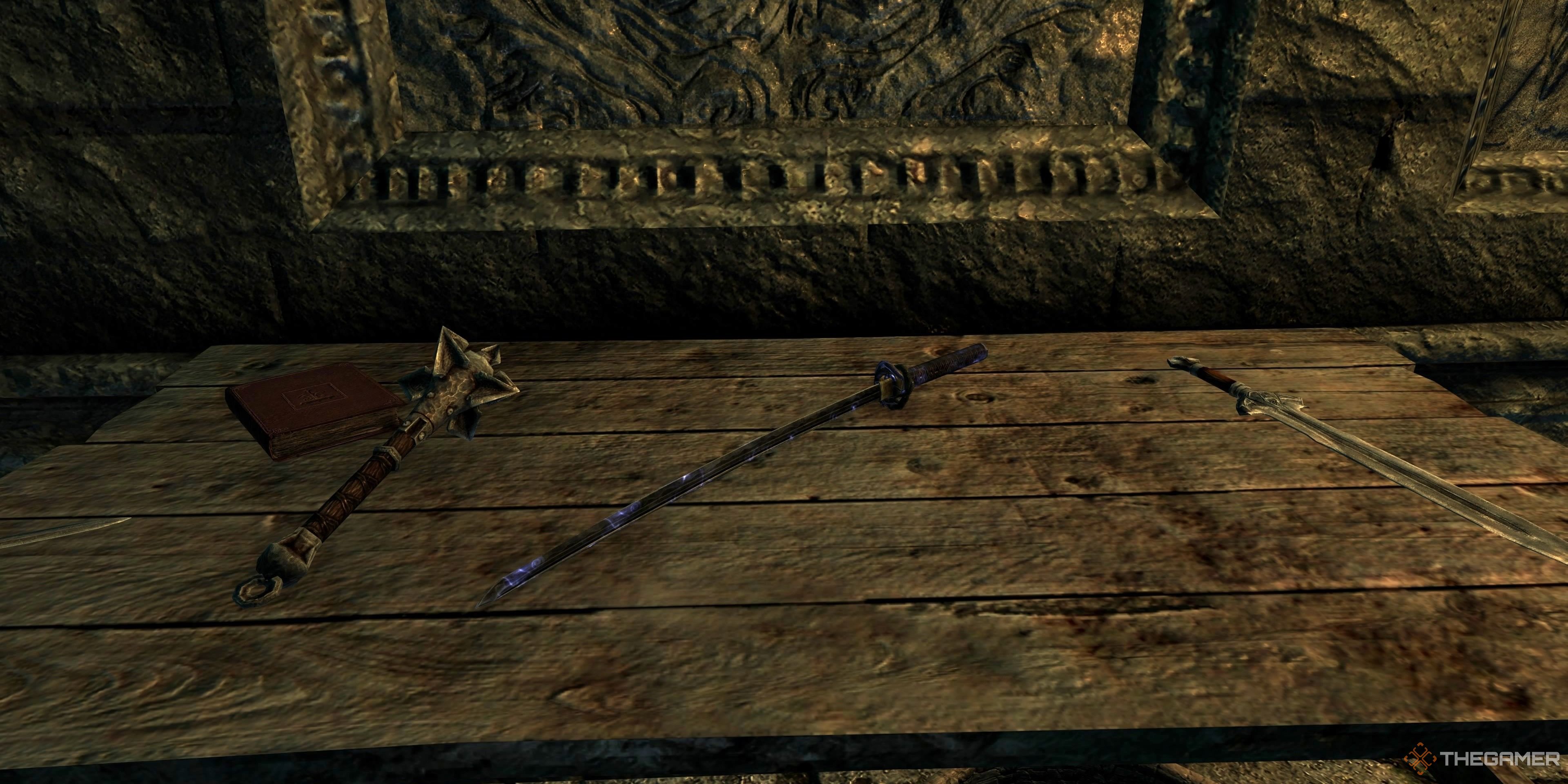 The image shows the Dragonbane laying on the table in Skyrim, there is an obvious blue glow on the edge/handle of the weapon and a mace and sword lay next to it on either side. The table is wooden with clear wear and tear over time.